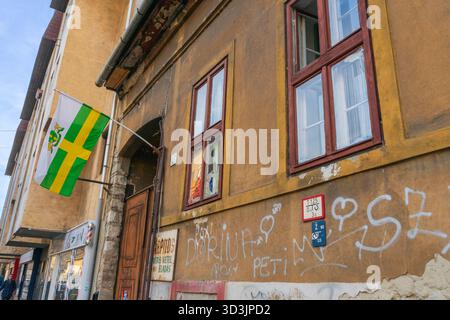 Budapester Fassade im 8. Bezirk mit Bezirksflagge, Graffiti und einem Kauf- und Verkaufsschild, das urbanen Schmutz und Straßenhandel einfängt. Stockfoto