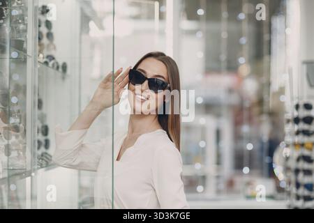 Nahaufnahme einer wunderschönen jungen Frau, die lächelt und an der Optikerecke im Einkaufszentrum Gläser pflückt und auswählt. Glückliche schöne Frau kaufen Augenbrillen Stockfoto