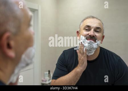 Porträt eines gutaussehenden grauhaarigen Mannes mittleren Alters, der Rasierschaum anwendet, um seinen Bart im Badezimmer zu trimmen Stockfoto