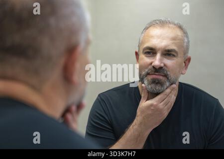 Ein gutaussehender Mann mittleren Alters, der in einem Badezimmer im Spiegel schaut und seinen Bart berührt Stockfoto