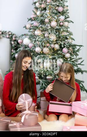 Zwei Mädchen eröffnen Geschenke vor einem geschmückten Weihnachtsbaum. Zwei Mädchen in roten Pullover sitzen auf einem weißen Teppich vor einem rosa und weiß verzierten Teppich Stockfoto