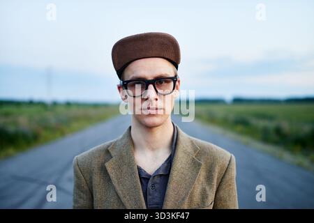 Porträt des New Age-Vertreters: Junger trendiger Mann in altmodischer Kleidung und Hornbrille, der in der Abenddämmerung mitten auf einer verlassenen Straße steht Stockfoto