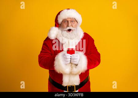 Der Weihnachtsmann in rotem Anzug mit weißem Bart hält ein Smartphone vor einem hellgelben Hintergrund für festliche Weihnachtszauber und Shopping-Vibes Stockfoto