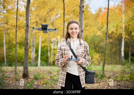 In einem lebhaften Wald, der mit Herbstfarben geschmückt ist, lächelt eine junge Frau, während sie geschickt eine Drohne steuert. Die Bäume um sie herum zeigen orange, gelbe und grüne Farbtöne. Stockfoto
