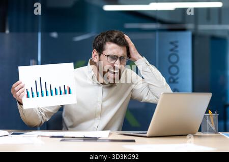 Schockierter junger Geschäftsmann, der am Tisch im Büro sitzt und Dokumente mit Grafiken auf dem Laptop-Bildschirm zeigt, und frustriert den Kopf hält. Stockfoto