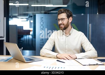 Ein lächelnder junger Mann sitzt mit Papieren im Büro und redet per Videoanruf auf einem Laptop. Stockfoto
