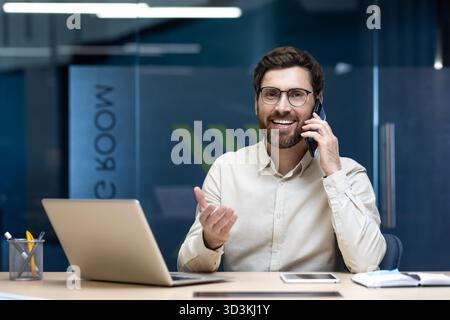 Porträt eines jungen, lächelnden und erfolgreichen Geschäftsmannes, der am Tisch im Büro sitzt, am Telefon spricht, mit der Hand Gesten zeigt und in die Kamera blickt. Stockfoto