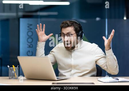 Angenehm überrascht, junger Mann mit Kopfhörern, der an einem Schreibtisch sitzt, begeistert auf den Laptop-Bildschirm starrt, die Hände hochhebt und sich über den Erfolg freut. Stockfoto