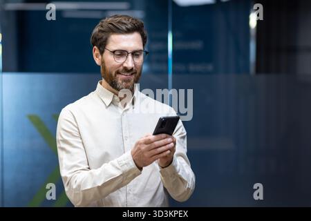 Lächelnder junger männlicher Büroangestellter, der drinnen steht und das Handy benutzt. Stockfoto