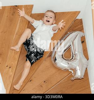 Oben ein Schuss von einem Jungen, der über Holzstufen liegt, mit ausgestreckten Armen neben einem silbernen Ballon mit 4 Bahnen. Verspielte, sorglose Komposition ideal zum Geburtstag Stockfoto