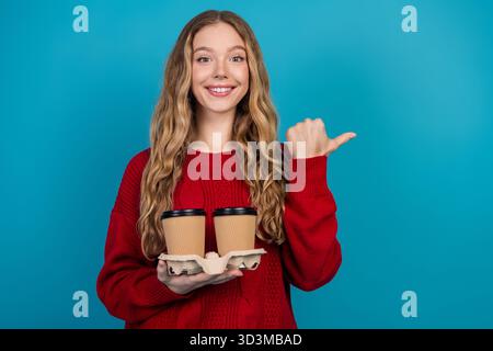 Eine junge Frau in einem roten Pullover, die zwei Kaffeetassen hält und eine fröhliche Geste vor einem hellblauen Hintergrund zeigt Stockfoto
