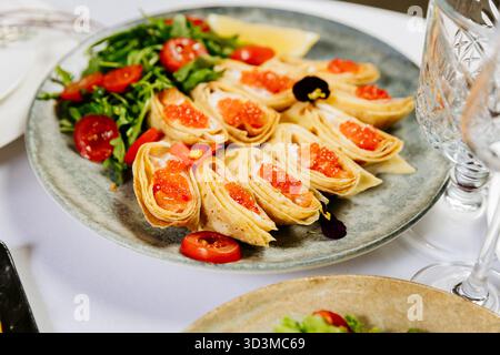 Festliche Krepprollen mit rotem Kaviar, Grün und Kirschtomaten auf elegantem Tisch Stockfoto