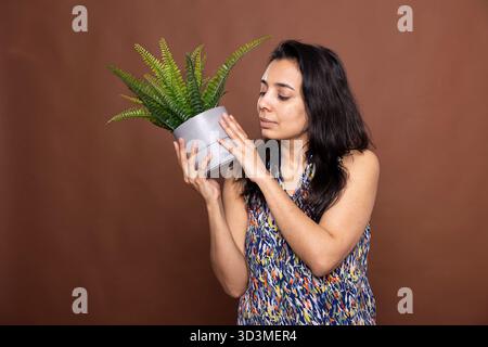 Indisches weibliches Modell mit heiterem Gesichtsausdruck hält Topfpflanzen und bewundert ihre Blätter. Eine ruhige, hübsche Frau, die die Schönheit der Gartenarbeit im Haus genießt, die Zimmerpflanze ergreift und ihr Grün schätzt. Stockfoto