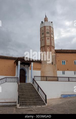 Die große Moschee von Chefchaouen, Marokko, mit ihrem einzigartigen achteckigen Minarett unter einem bewölkten Himmel. Steintreppen führen zu einem dekorativen Holzeingang Stockfoto