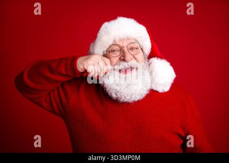 Der Weihnachtsmann in einem roten Pullover und Hut lächelt vor einem auffälligen roten Hintergrund warm und bereit für die Weihnachtszeit Stockfoto