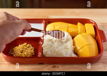 Eine Hand greift nach einem Löffel Sirup, der bereit ist, über eine köstliche Portion süßen Kleberreis, frische Mangoblätter und knusprige Mungbohnen hineinzuträufeln Stockfoto