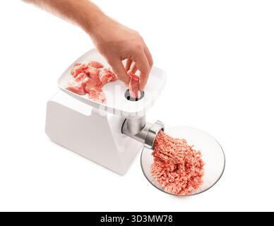 Die Hand einer Person schiebt ein rohes Stück Schweinefleisch in eine weiße elektrische Fleischwolf, wobei sich das Hackfleisch in einer Glasschale sammelt. Hochwertige Fotos Stockfoto