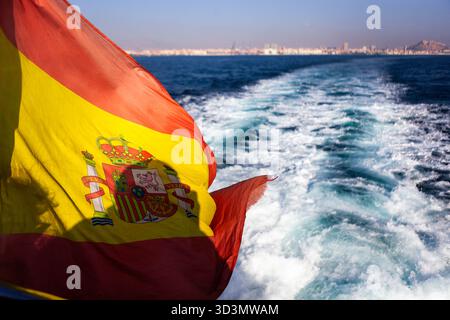 Eine spanische Flagge flattert auf einer Fähre nach Tabarca Island, nahe Alicante. Die Aussicht von der Rückseite des Bootes. Die Stadt am Horizont. Stockfoto