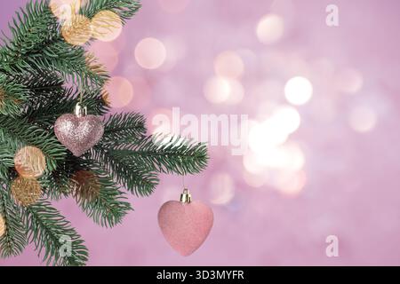 Weihnachtsbaumzweige mit herzförmigen Kugeln vor verschwommenem Licht auf rosa Hintergrund, Bokeh-Effekt Stockfoto