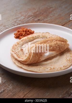 indische Dosa mit roter Kokos Sambol oder Chutney auf weißem Teller vor rustikalem Holztisch, südindische Küche und traditionelles Frühstück Stockfoto