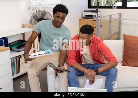 Bei der Überprüfung von Tablet- und Designmodellen sitzen verschiedene männliche Kollegen auf Sofa im Büro, mit Drucker Stockfoto
