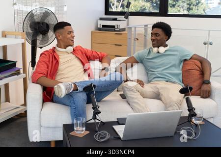 Aufnahme verschiedener männlicher Freunde, die Podcasts auf hellgrauem Sofa im Studio mit Mikrofonen halten Stockfoto