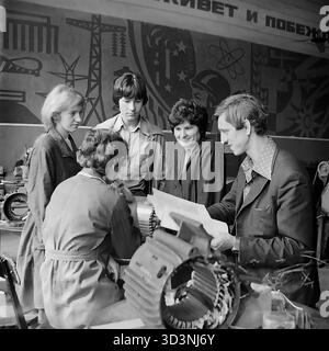 Ein Archivfoto aus dem Jahr 1981 zeigt ein Produktionstreffen in der AIZ (Armature-Isolator Plant) in Sloviansk, ukrainische SSR. Ein männlicher Ingenieur oder Vorarbeiter hält eine technische Zeichnung und leitet ein Team von vier weiblichen Arbeitern ein. Sie sind um ein großes Industriekomponent, möglicherweise den Stator eines Elektromotors, angeordnet. Die Szene entfaltet sich in der Werkstatt, direkt vor einem großen, ikonischen sowjetischen Propagandabild, das Industrie, Wissenschaft (Atome) und Fortschritt darstellt. Dies fängt industrielle Arbeit und ideologische visuelle Kultur im friedlichen Donbass vor dem Krieg ein Stockfoto