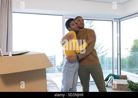 Ein vielseitiges Paar, das in einem modernen Apartment steht, mit Umzugskartons, Laptop und zusammengefalteter Decke Stockfoto