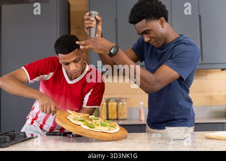 Verschiedene männliche Freunde bereiten Pizza in der modernen Küche zu, mit Holzpizzaschalen und Pfeffermühle Stockfoto