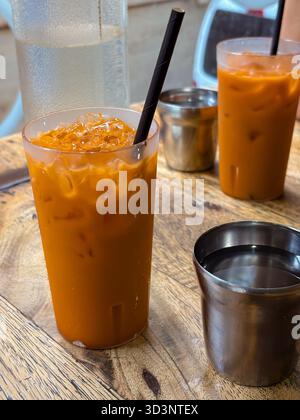 Thai-Eistee in klaren Plastikbechern mit Eis und schwarzen Strohhalmen sowie Wassergläsern auf einem rustikalen Holztisch in ungezwungener Atmosphäre. Stockfoto