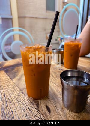 Ein eiskaltes thailändisches Teegetränk mit schwarzem Strohhalm sitzt auf einem rustikalen Holztisch in einem Café. Ein weiterer Drink und Wasserbecher sind im Hintergrund. Stockfoto