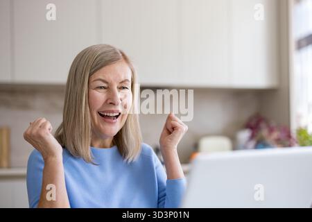 Die Seniorin feiert ihren Erfolg und ihr Erreichen von Zielen, während sie gute Nachrichten erhält oder online mit einem Laptop zu Hause gewinnt, und drückt Freude und Begeisterung aus Stockfoto
