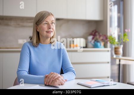 Die Seniorin sitzt an einem weißen Tisch in ihrer modernen Küche, mit gefesselten Händen, blickt nachdenklich weg, während sie ihre zukünftigen Ziele und täglichen Aufgaben in einem offenen Notizbuch plant Stockfoto