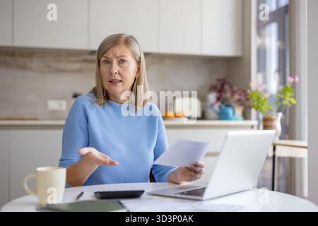 Seniorin in der Küche sah gestresst und verwirrt aus, während sie Rechnungen, Laptops und Papierkram überprüfte, mit Haushaltsbudget, Finanzen, Rente und steigenden Ausgaben zu kämpfen hatte Stockfoto