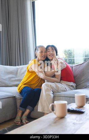 Lächelnde ältere asiatische Mutter und Erwachsene Tochter, die sich auf dem Sofa im Wohnzimmer umarmen, mit Kaffeetassen Stockfoto