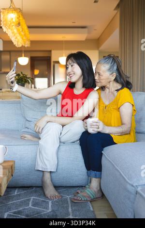 Lächelnde asiatisch-chinesische Großmutter und Enkelin machen Selfie auf dem Sofa, mit Smartphone und Becher Stockfoto