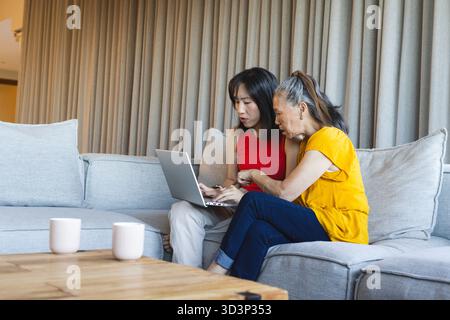Aussehende asiatisch-chinesische Mutter und Tochter sehen zu Hause einen silbernen Laptop auf dem Sofa, mit Kaffeetassen Stockfoto