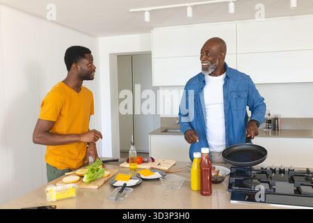 Kochen von afroamerikanischen Vater und Sohn, schneiden Salat und arrangieren Burgerbrötchen auf der Kücheninsel Stockfoto