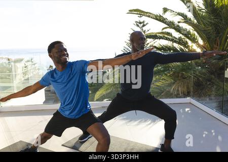 Üben Sie afroamerikanische Männer, die Warrior II auf einer gefliesten Außenterrasse mit Yogamatten spielen Stockfoto