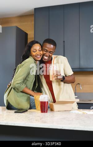 Afroamerikaner umarmt, lacht auf Kücheninsel, hält kalte Getränke mit Take-out-Tasche Stockfoto