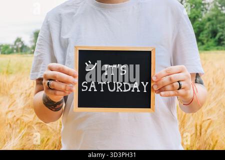 Eine Person, die ein Schwarzes Brett hält, auf dem die Worte „Samstag“ stehen. Die Person lächelt, was auf eine positive und glückliche Stimmung hindeutet Stockfoto