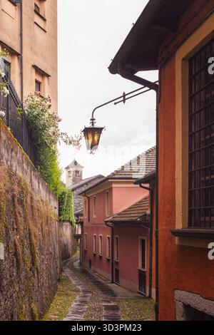 Malerische, enge Gasse auf der Insel San Giulio mit pastellfarbenen Wänden und Vintage-Laterne, Kopfsteinpflasterweg, der sich zwischen alten Häusern in Richtung einer Steinglocke schlängelt Stockfoto
