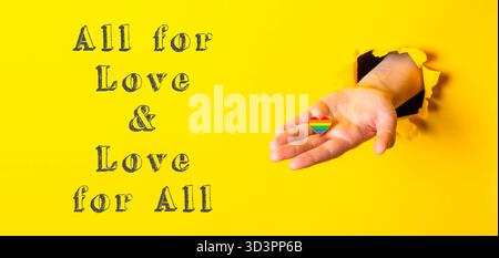 Eine Hand hält ein Regenbogenherz mit den Worten „alle für Liebe“ und „Liebe für alle“ darunter Stockfoto