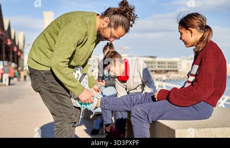 Ein Mann hilft einem Mädchen, ihre Schuhe anzuziehen, während zwei Jungs in der Nähe sitzen. Sie sind an einem sonnigen Tag in der Nähe des Ufers bereit für lustige gemeinsame Aktivitäten. Stockfoto