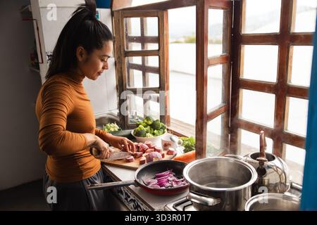 Eine Frau kocht in einer ecuadorianischen Küche, schneidet Gemüse auf einem Holzbrett an einem sonnigen Fenster. Es gibt Töpfe und frische Zutaten, die ein widerspiegeln Stockfoto