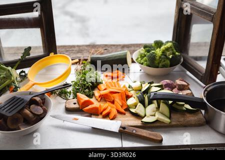Eine Vielzahl von frischem Gemüse, einschließlich Karotten, Brokkoli und Zucchini, sind kunstvoll auf einem hölzernen Schneidebrett in einer rustikalen Küche angeordnet Stockfoto