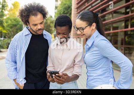 Ein vielseitiges Business-Team aus drei Fachleuten, das sich mit einem Lächeln beschäftigt hat, während es Informationen auf einem Smartphone in einer urbanen Umgebung im Freien überprüft und präsentiert Stockfoto