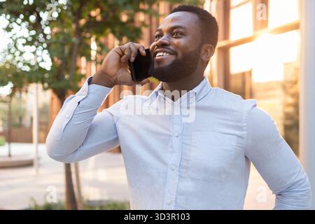 Selbstbewusster Geschäftsmann in einem lässigen weißen Hemd, lächelnd während eines Telefongesprächs im Freien, umgeben von urbaner Architektur und Stockfoto