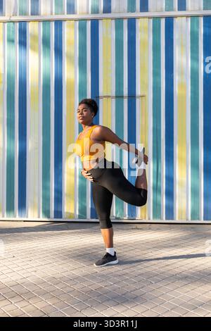 Eine junge Lateinfrau streckt ihr Bein, während sie vor einer lebendigen gestreiften Wand steht. In sportlicher Kleidung bereitet sie sich auf einen Lauf unter der Mo vor Stockfoto
