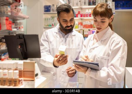 Zwei Apotheker in weißen Labormänteln stehen in einer Apotheke und besprechen aufmerksam die Medikamentendetails, während einer auf eine Tablette schreibt und der andere ein p hält Stockfoto